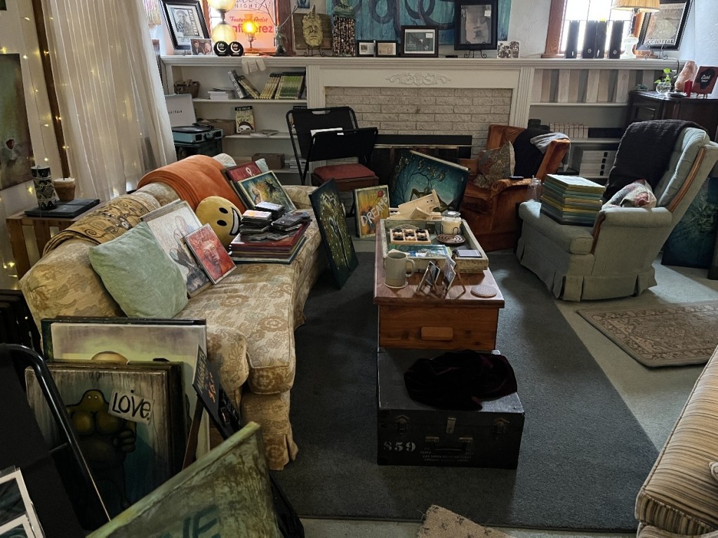 A cozy living room cluttered with various paintings, framed art, and stacks of art supplies on the furniture and floor, depicting the vibrant chaos of a creative home during the holiday season.