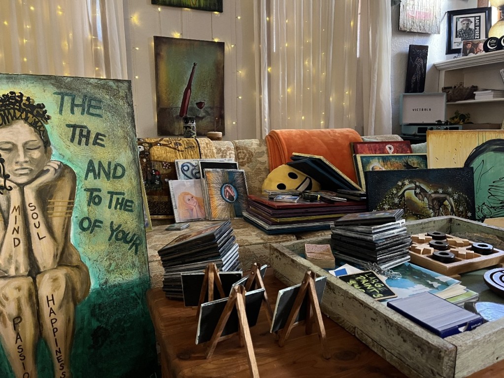 A cluttered living room showcasing an array of paintings, art supplies, and framed artworks. A large canvas with a figure is prominently displayed, surrounded by stacks of smaller prints, notebooks, and colorful throws. Warm lighting enhances the creative chaos present in the space.