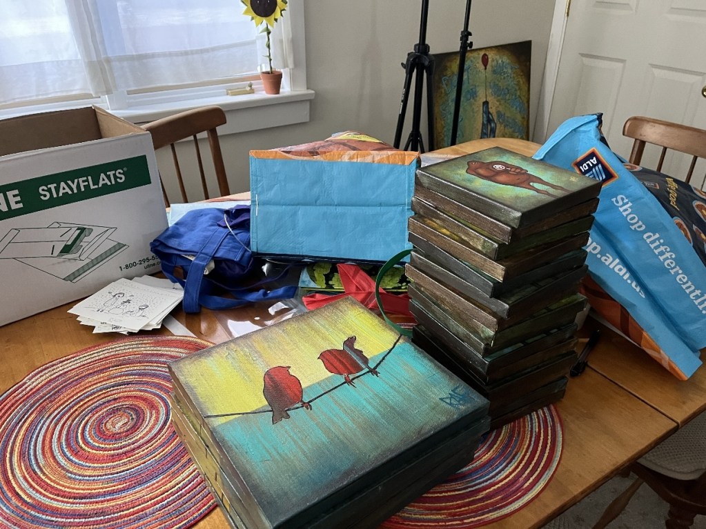 A cluttered dining table filled with stacks of paintings, packaging materials, reusable tote bags, and an open shipping box, creating a vibrant and chaotic creative workspace.