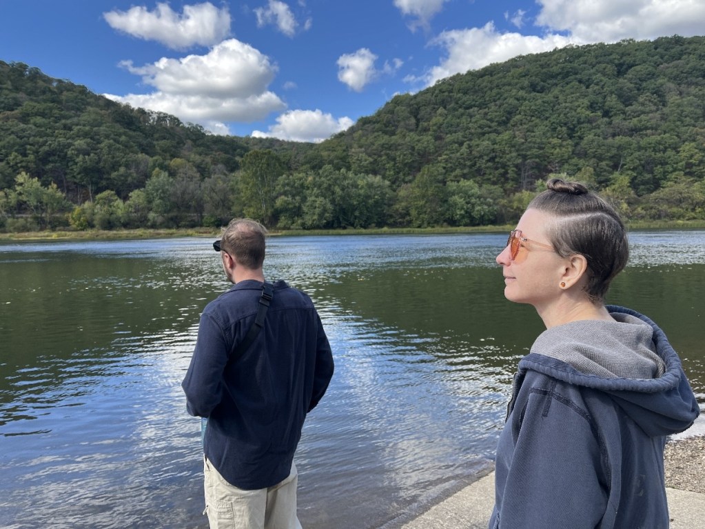 A man and a woman standing by a calm river, looking out at the water with green hills in the background and a partly cloudy sky.