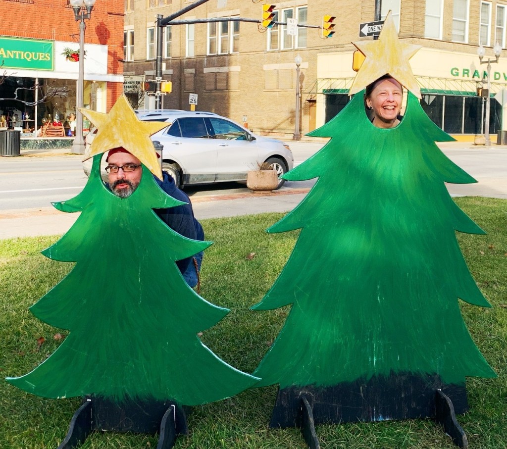 Two people playfully posing in cutouts shaped like Christmas trees, with a star on top, smiling against a festive street background.