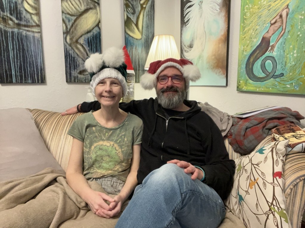 Two people sitting on a couch in a cozy living room, wearing festive hats. The room features colorful artwork on the walls and a warm lamp, creating a welcoming atmosphere.