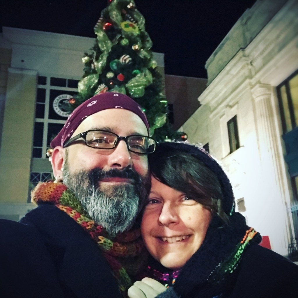 A smiling couple wearing winter scarves and hats, standing in front of a decorated Christmas tree at night.