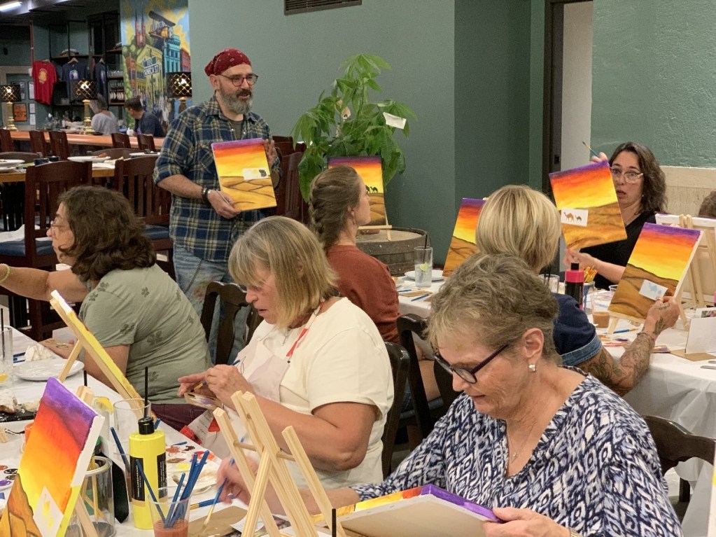 A group of people engaged in a painting class, with a male instructor assisting. Participants are holding their artworks featuring sunset scenes.