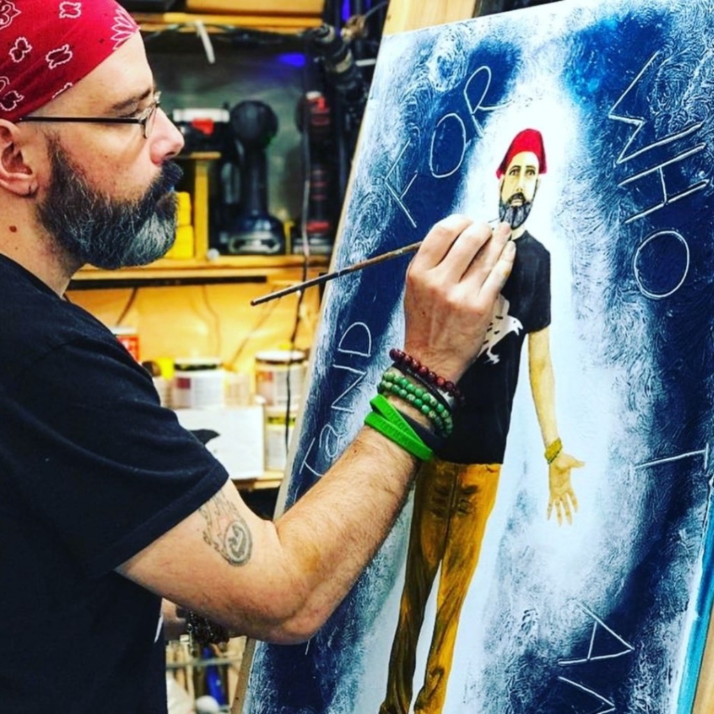 An artist is painting on a canvas in a workshop, wearing a red bandana and glasses, surrounded by art supplies.