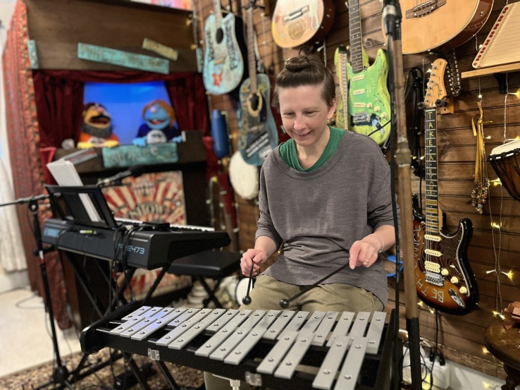 A person playing a xylophone in a music studio filled with various instruments, including guitars and puppets in the background.