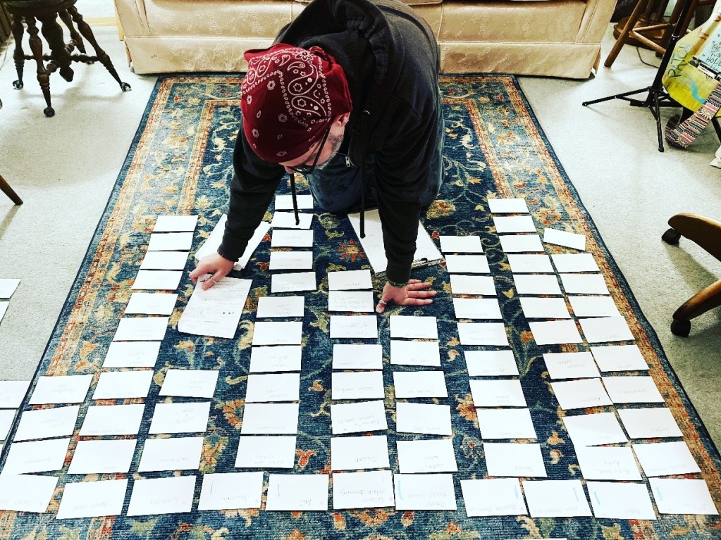 A person kneeling on a carpet, organizing a variety of index cards spread out in rows on the floor, while surrounded by furniture.