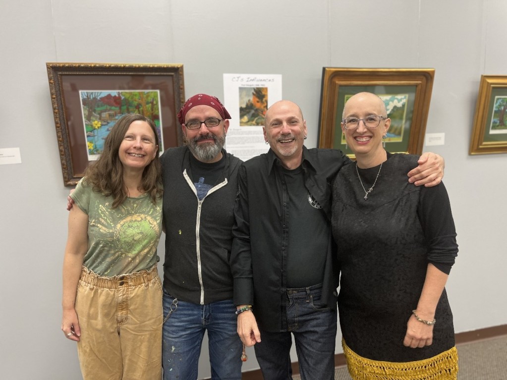 A group of four people posing together in an art gallery, smiling and standing in front of paintings. They are wearing casual clothing and appear to be enjoying the event.