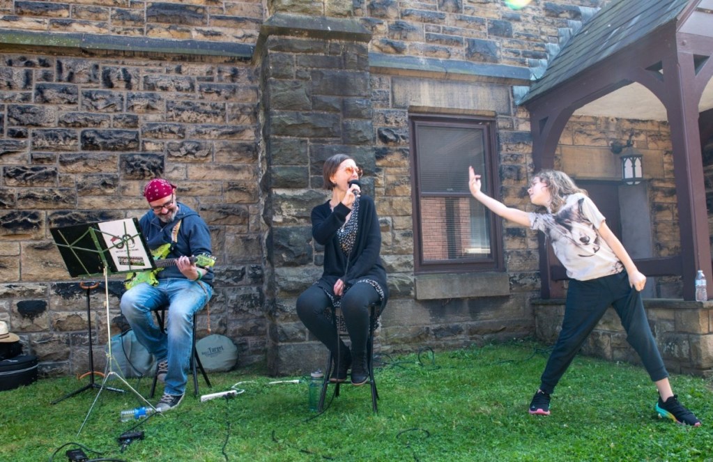 A musician playing guitar on stage, a vocalist singing into a microphone, and a child dancing expressively in front of them, all set against a stone wall backdrop at an outdoor event.