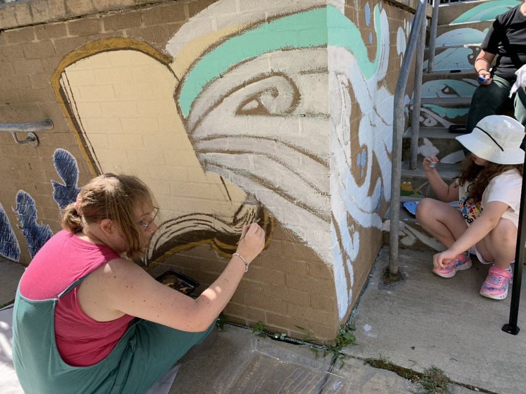Artists painting a mural of an open book on the wall of the Oil City Library entrance, with children actively participating in the creative process.