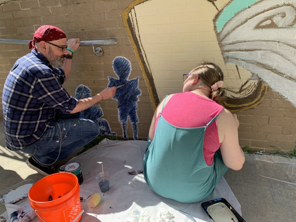 Artists painting a mural at the entrance of a library, focusing on children's silhouettes and a large open book, capturing creativity and community engagement.