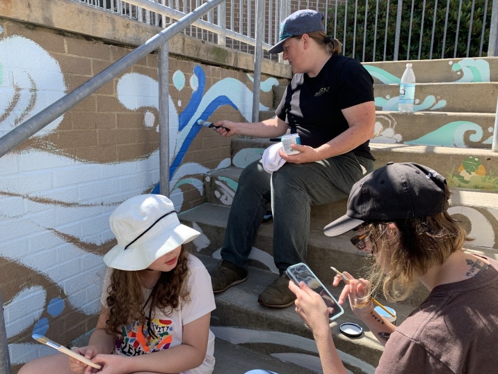 Three individuals working on a mural project at the Oil City Library, with one person painting on the steps and two others observing and discussing.