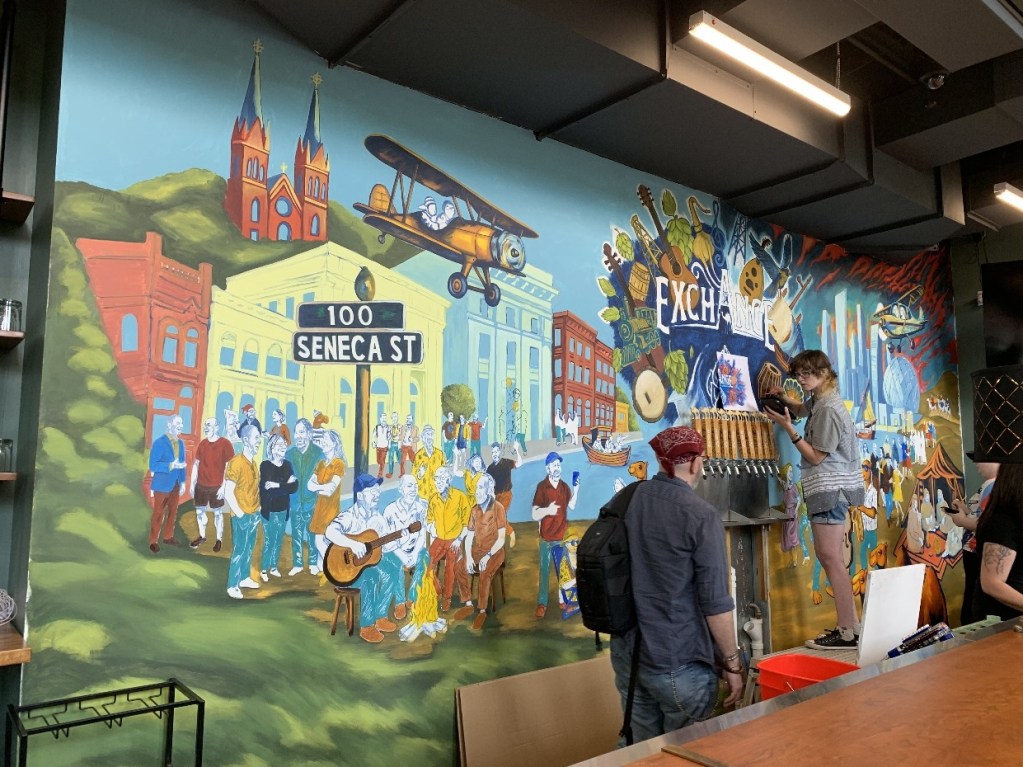 A colorful mural depicting a scene at 100 Seneca Street in Oil City, featuring various characters, buildings, and elements representing the local culture and history.