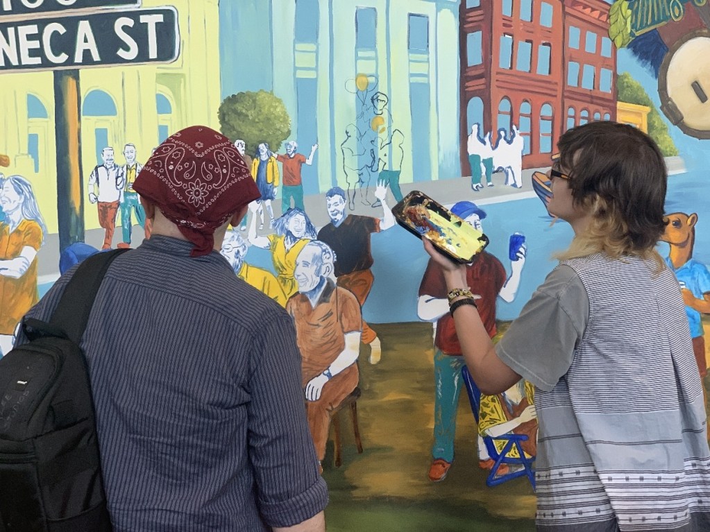 Two artists are working on a colorful mural depicting scenes and portraits of people in Oil City. One artist is wearing a bandana and holding a paint palette, while the other is observing the mural.