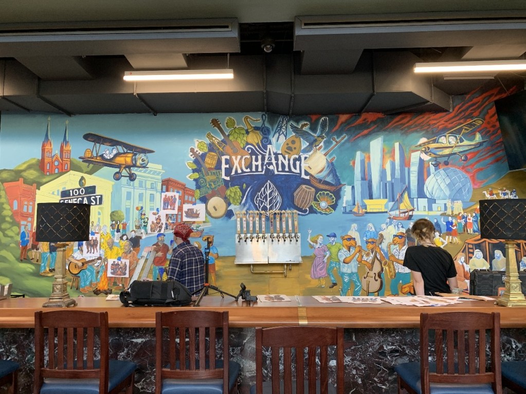 A vibrant mural at The Exchange in Oil City, featuring colorful depictions of local landmarks, community members, and cultural elements, with two artists working on the piece.
