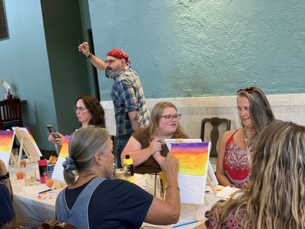 A lively paint and sip event at The Exchange with participants painting on easels, enjoying drinks, and engaging with each other in a creative atmosphere.
