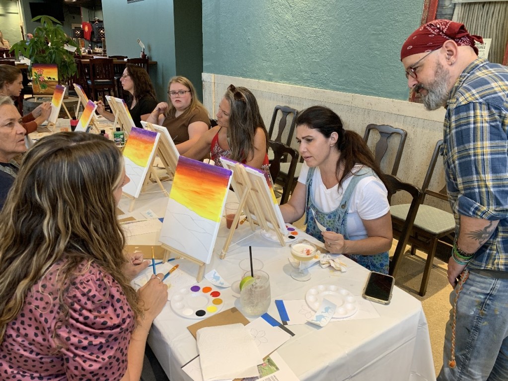 A group of people engaged in a paint and sip event, with canvases set up in front of them and various painting supplies on the table. The atmosphere appears lively and creative as participants enjoy the experience.