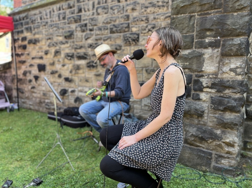A woman singing into a microphone while seated, accompanied by a man playing a small instrument in the background. They are performing outdoors near a stone wall.