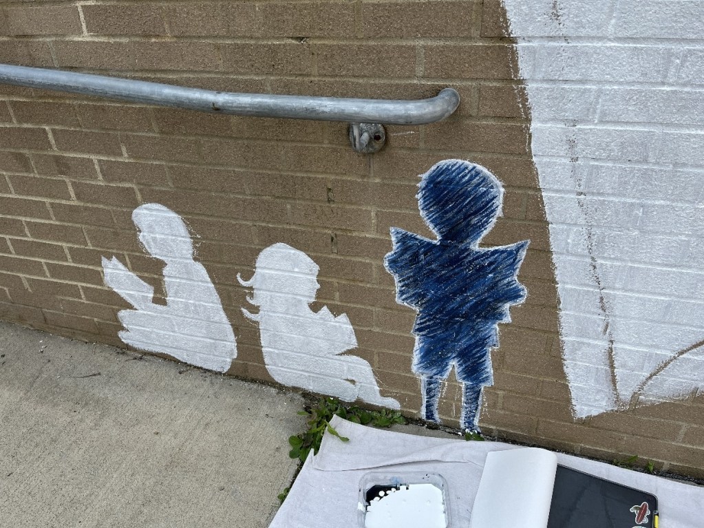 Silhouettes of children reading on a brick wall, with a painted railing above them, showcasing early stages of a mural at the Oil City Library.
