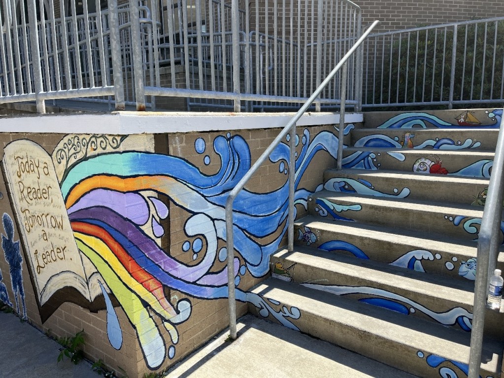 A mural on the entrance wall and stairs of the Oil City Library, featuring an open book with the phrase 'Today a Reader, Tomorrow a Leader.' Colorful waves and sea creatures flow from the book, creating an engaging and whimsical environment for children.