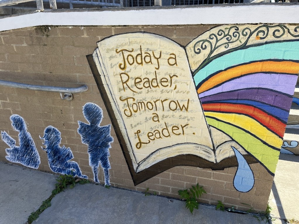 A colorful mural at the entrance of the children's section of the Oil City Library, featuring a large open book with the words, 'Today a Reader, Tomorrow a Leader.' Vibrant waves and a rainbow emerge from the book, alongside silhouettes of children interacting with the space.