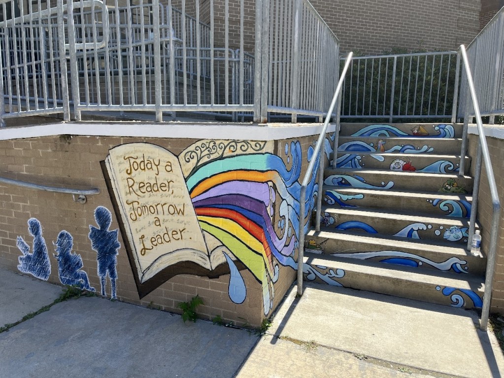 A colorful mural at the Oil City Library featuring a large open book with the quote 'Today a Reader, Tomorrow a Leader,' vibrant waves and rainbow colors, and playful sea creatures, adorning the entrance to the children's section.