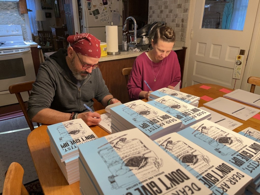 Two individuals are seated at a table covered with stacks of books titled 'Dear Artist, Don't Give Up!' as they sign copies. One person wears a red bandana and glasses, while the other has their hair in a bun and wears a purple top.