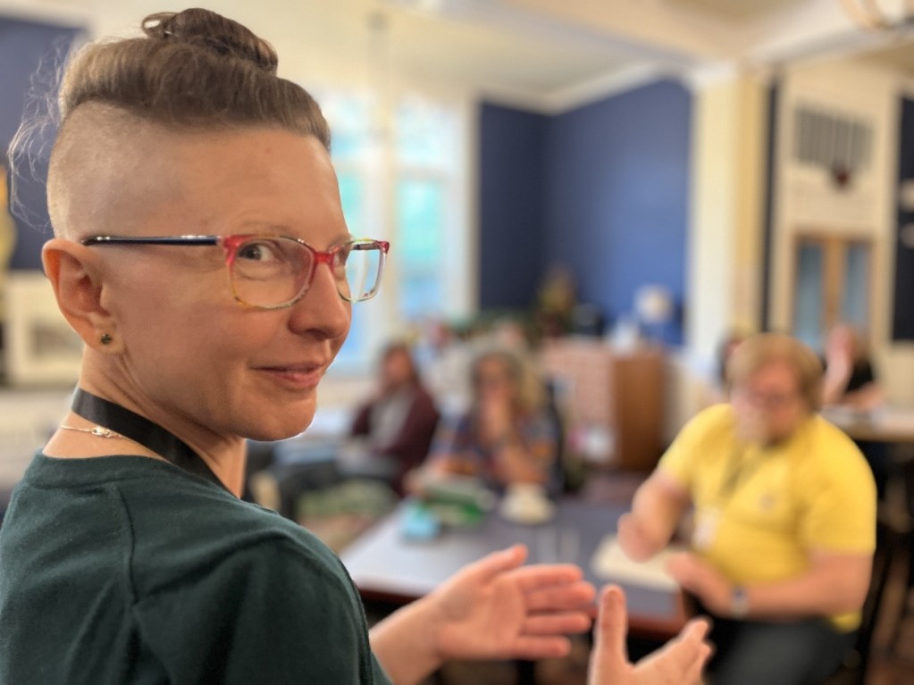 A person with short hair and glasses gestures while speaking to an audience in a workshop setting, with attendees seated at tables in the background.