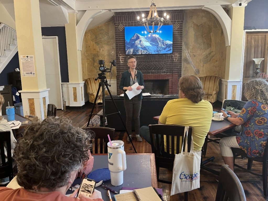 A speaker stands in front of an audience in a cozy venue, discussing topics related to creativity and overcoming challenges for artists, while a presentation is displayed on a screen behind.