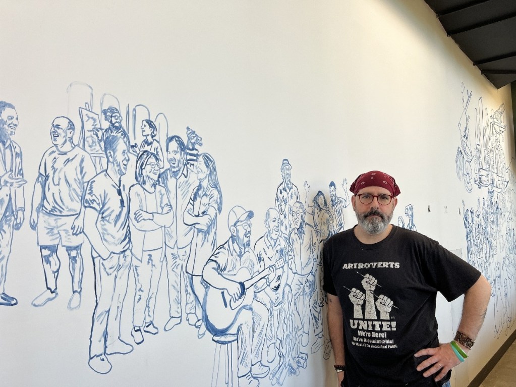 A mural depicting a diverse group of people in various poses and expressions, with an artist standing in front of it. The mural features a light blue outline style, capturing the essence of the local community.