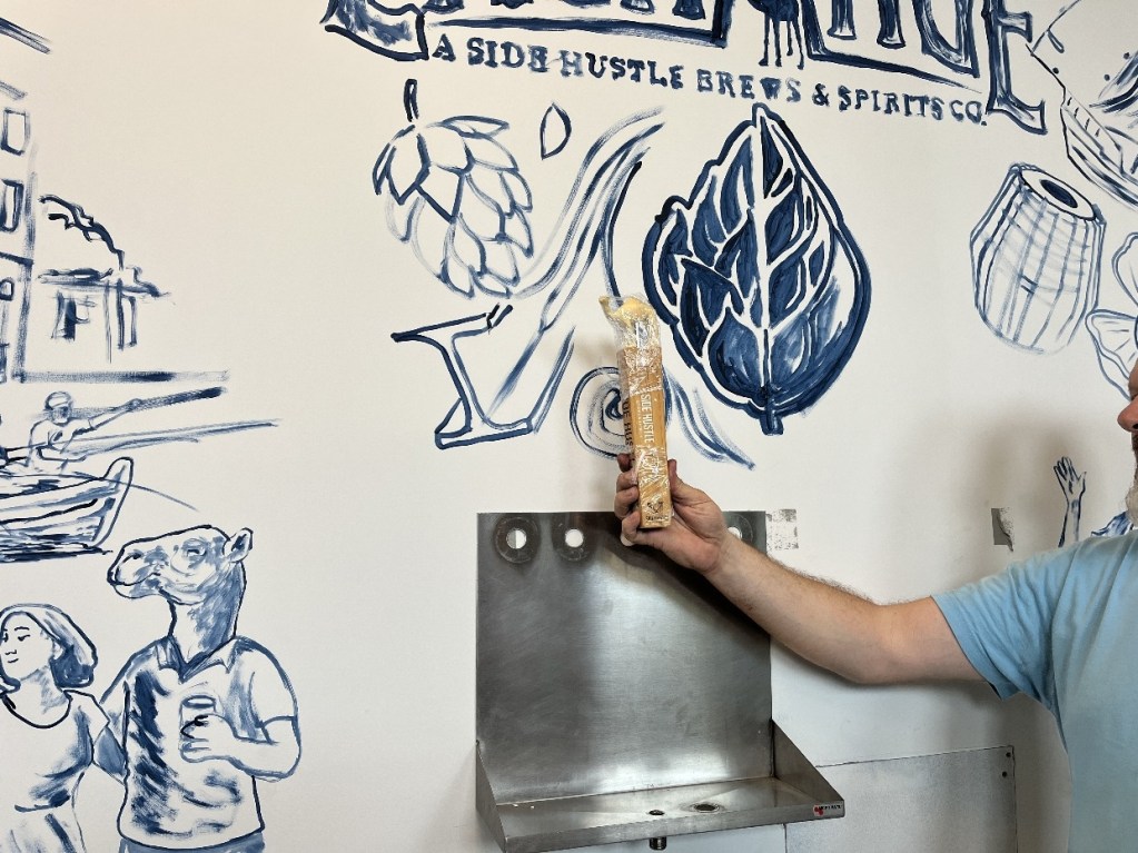 A person holding a snack while standing in front of a partially painted mural depicting various elements related to Side Hustle Brews & Spirits, including a camel, hops, and a river scene.