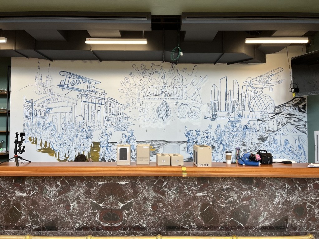 A mural featuring a lively scene with various characters and historical elements, depicting the vibrancy of Oil City, on the wall behind a wooden bar at The Exchange.