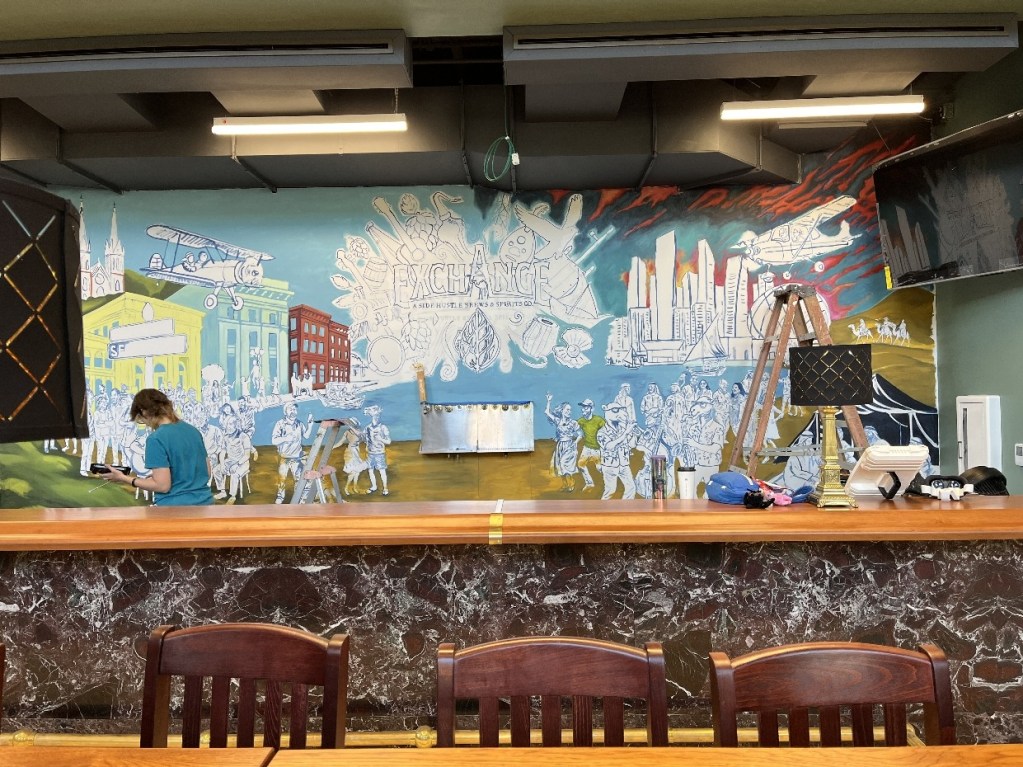 A mural in progress showcasing vibrant scenes and characters representing the history of Oil City, with an artist painting on the wall while ladders and tools are visible.