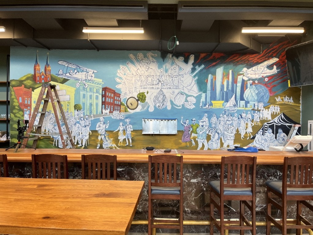 A partially completed mural in The Exchange, Oil City, featuring iconic local scenes, historical buildings, and numerous people depicted in a vibrant artistic style, with a wooden bar and ladder in the foreground.