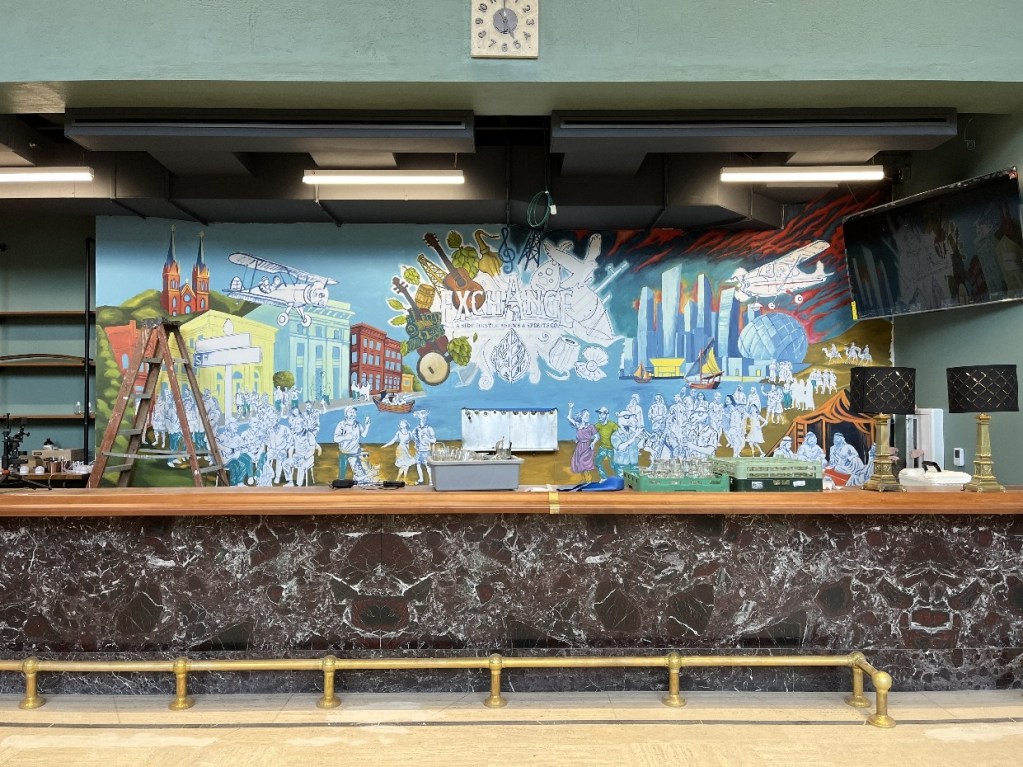 A vibrant mural at The Exchange in Oil City, depicting a mix of historical and contemporary elements, featuring landmarks, people, and cultural symbols, with a wooden bar in the foreground.