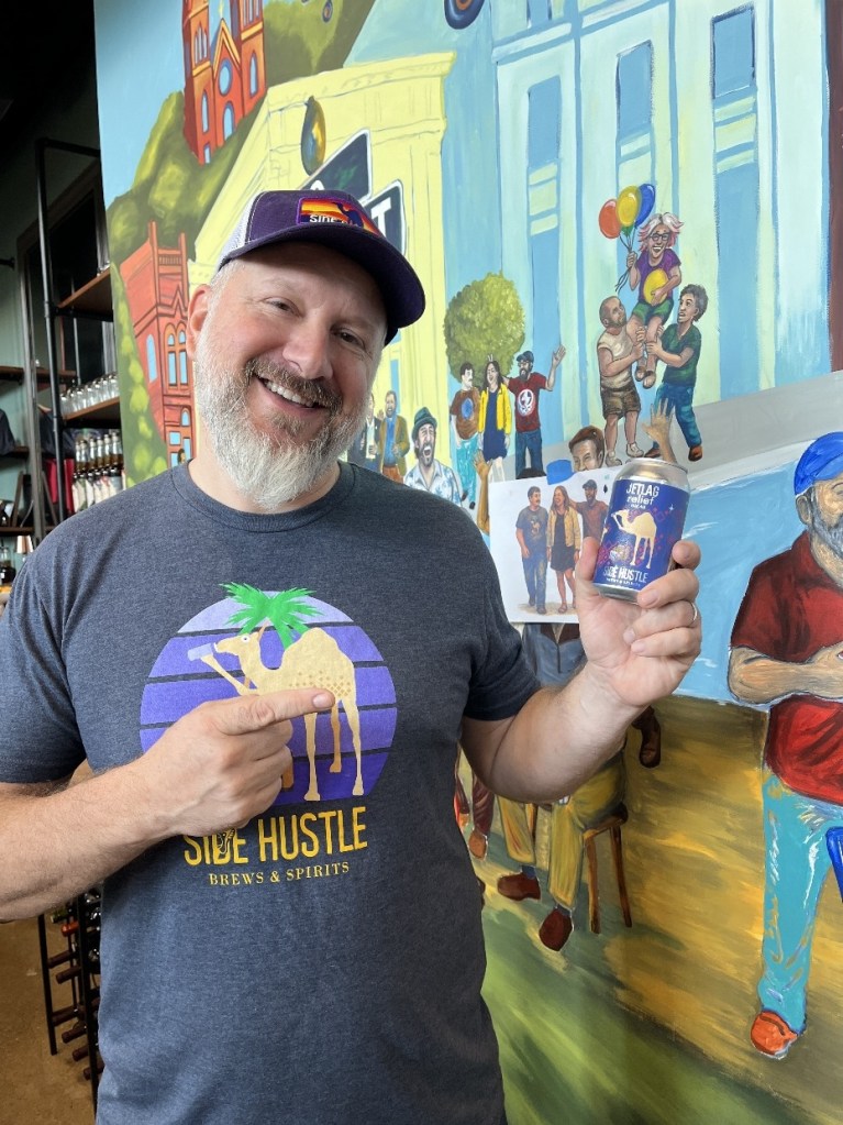 A man wearing a 'Side Hustle Brews & Spirits' shirt smiles while holding a can of beer in front of a colorful mural depicting various people and scenes associated with the local community.