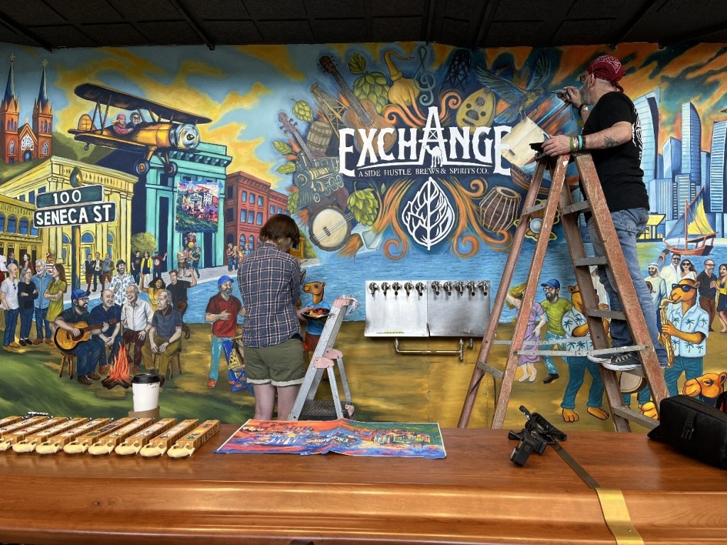 Two artists painting a mural in The Exchange, Oil City, featuring vibrant scenes of community life, historic buildings, and cultural elements. One artist works on a ladder while the other paints on the ground, surrounded by musical instruments and the mural's colorful design.
