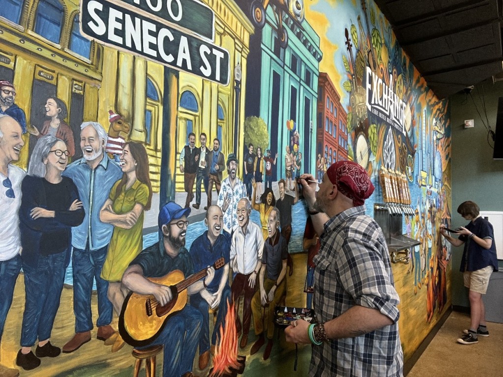 An artist painting a vibrant mural at The Exchange in Oil City, featuring various people and a prominent street sign.