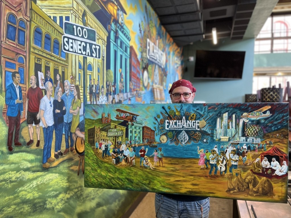 A person holding a large painted mural in front of a colorful backdrop depicting a lively street scene at 100 Seneca Street, featuring various figures and elements representing the community of Oil City.