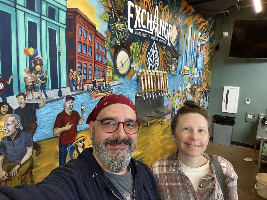 Selfie of two artists in front of a colorful mural depicting community scenes at The Exchange in Oil City.