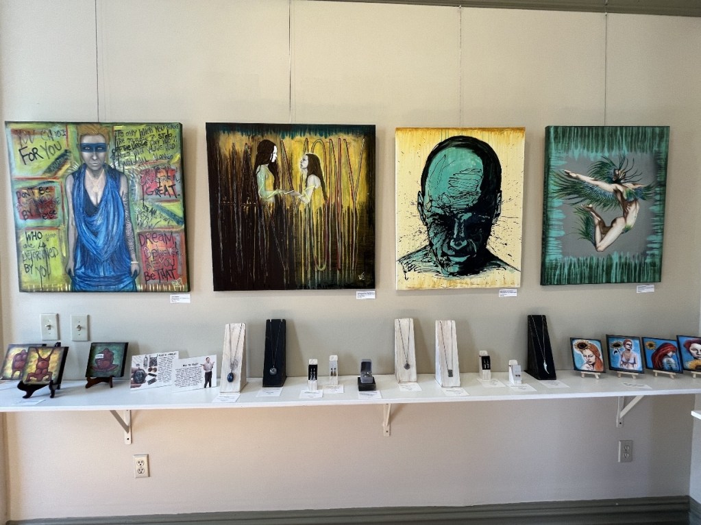 A display of various artworks at the Red Brick Gallery, featuring vibrant paintings and intricate jewelry pieces, showcasing a blend of artistic styles and creativity.