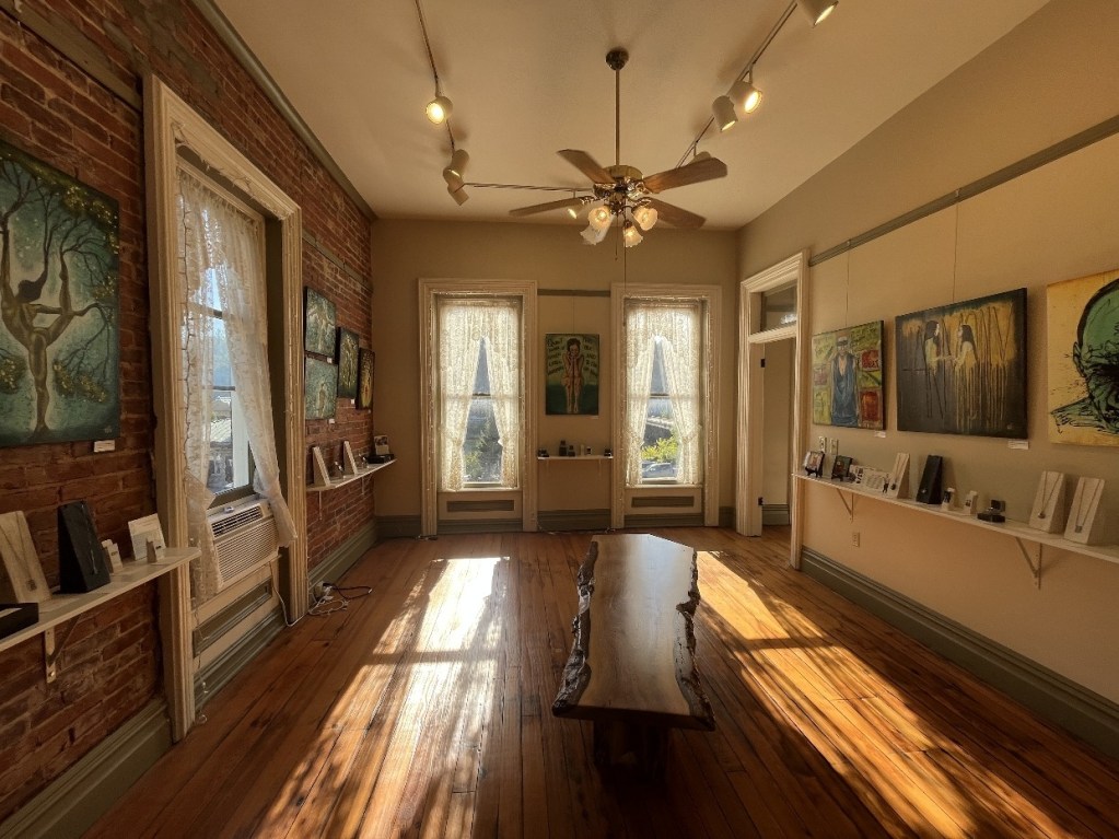 Interior view of the Red Brick Gallery in Foxburg, PA, featuring artwork displayed on the walls and shelves, with natural light illuminating the wooden floor.