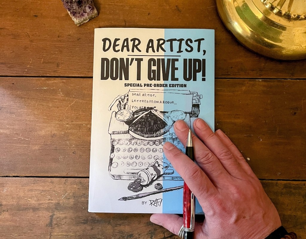 A hand holding a pencil over the cover of the book 'Dear Artist, Don't Give Up!' which features a typewriter illustration and text indicating it is a special pre-order edition.