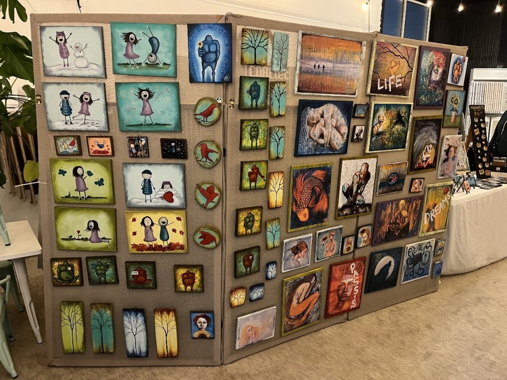 A display wall featuring a variety of colorful paintings and artworks, showcasing different themes and styles, set up at a local pop-up shop.