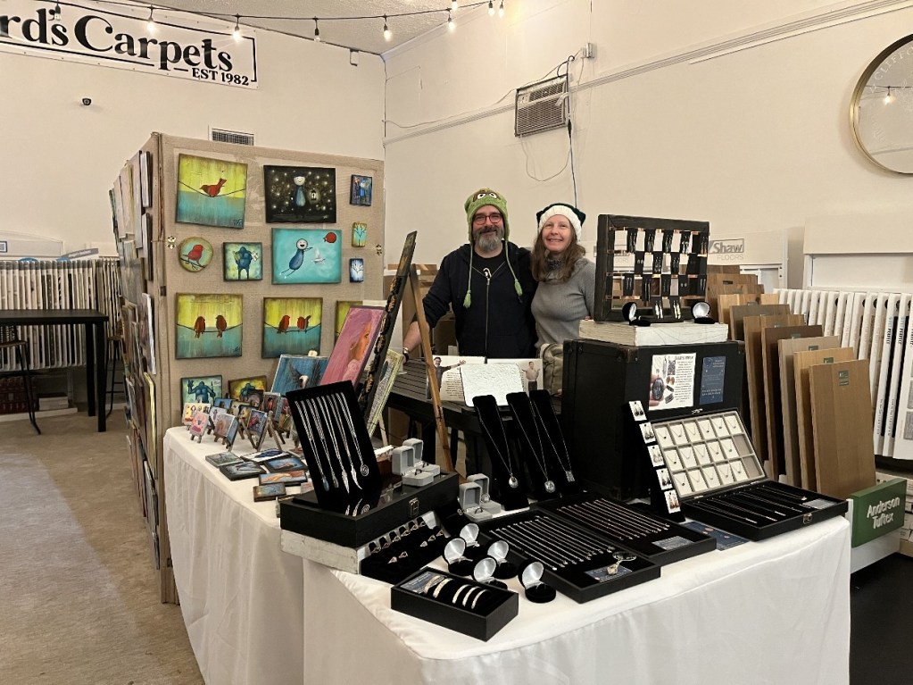 A couple stands behind a vendor booth at a craft fair, surrounded by colorful artwork and handcrafted jewelry. The booth features a display of paintings, photographs, and various jewelry items.