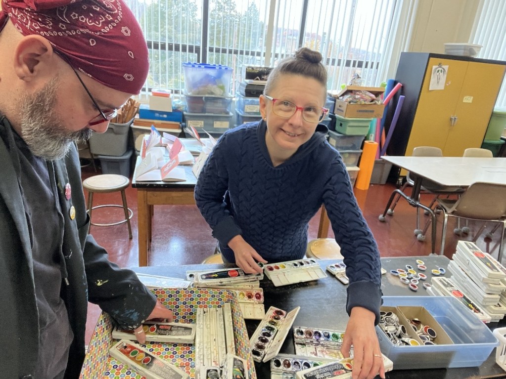 Two instructors preparing art supplies in a classroom setting, surrounded by various art materials and a cheerful atmosphere.