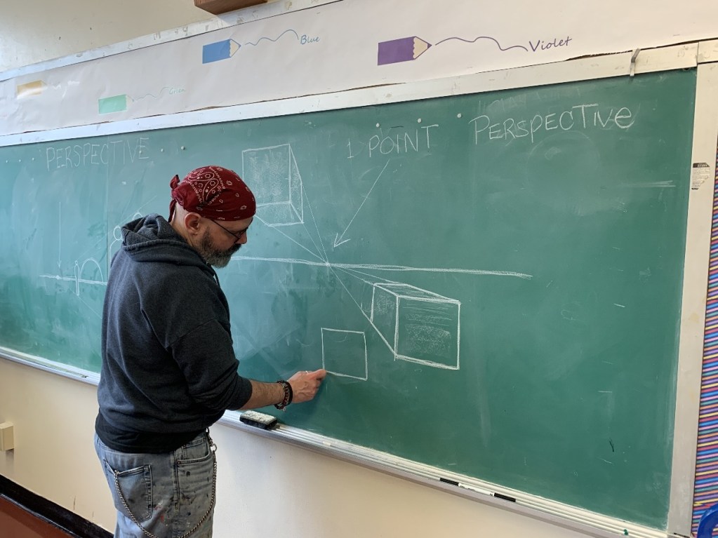 An instructor demonstrating one-point perspective drawing techniques on a chalkboard in a classroom setting.