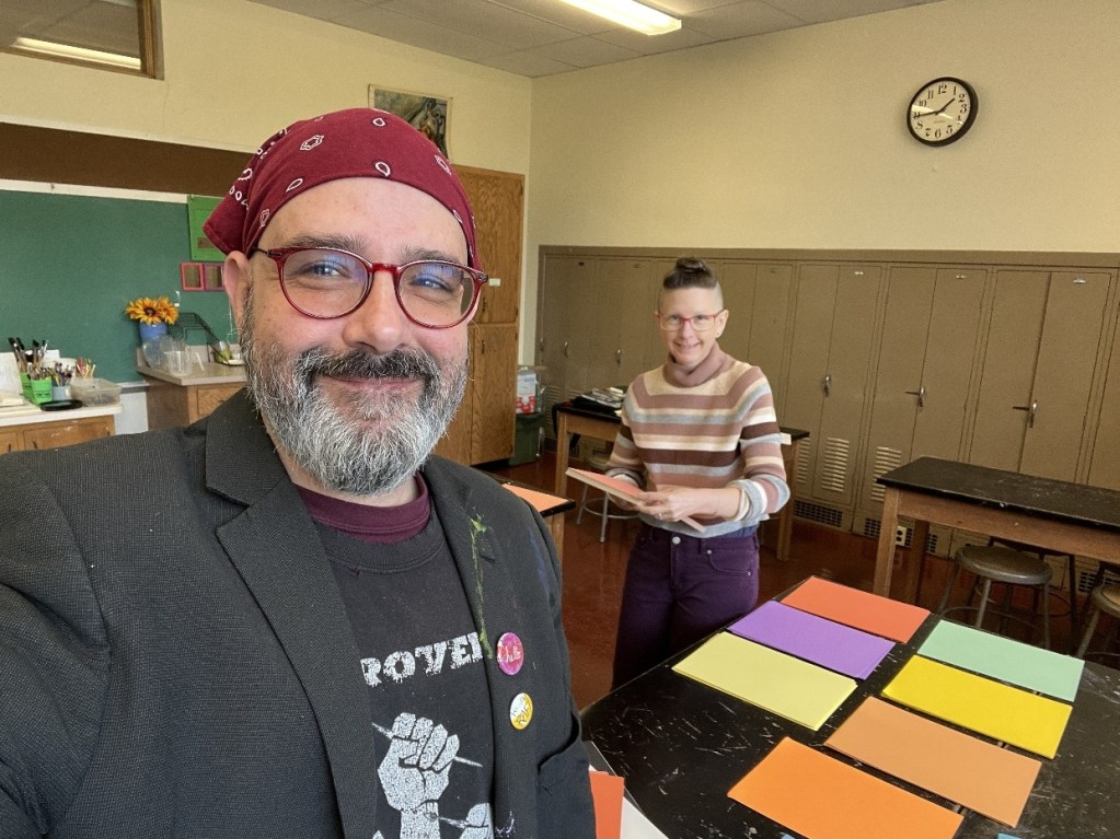 Two art instructors smiling in a classroom, with colorful papers spread across a table and art supplies in the background.
