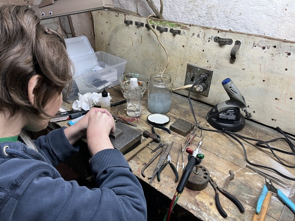 A person with short hair is focused on a workbench, using tools and materials for a creative project, surrounded by various art supplies and equipment.
