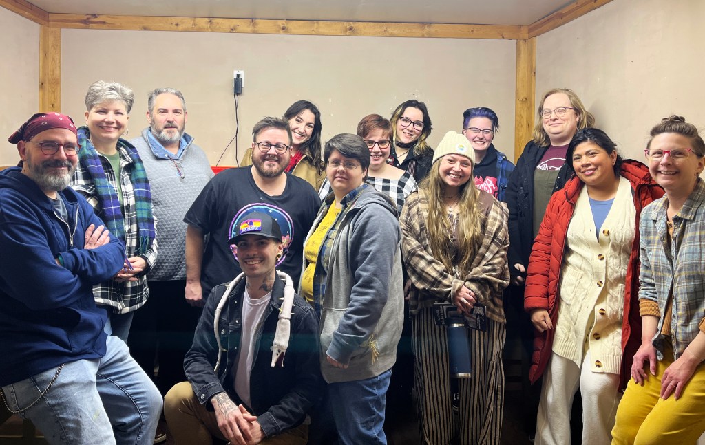 A diverse group of fifteen people smiling and posing together in a room with wooden accents and neutral walls.